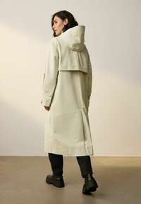 Kvinna som bär en beige lång huvad trenchcoat, svarta byxor och svarta kraftiga stövlar, står på ett slätt golv mot en enkel vägg.