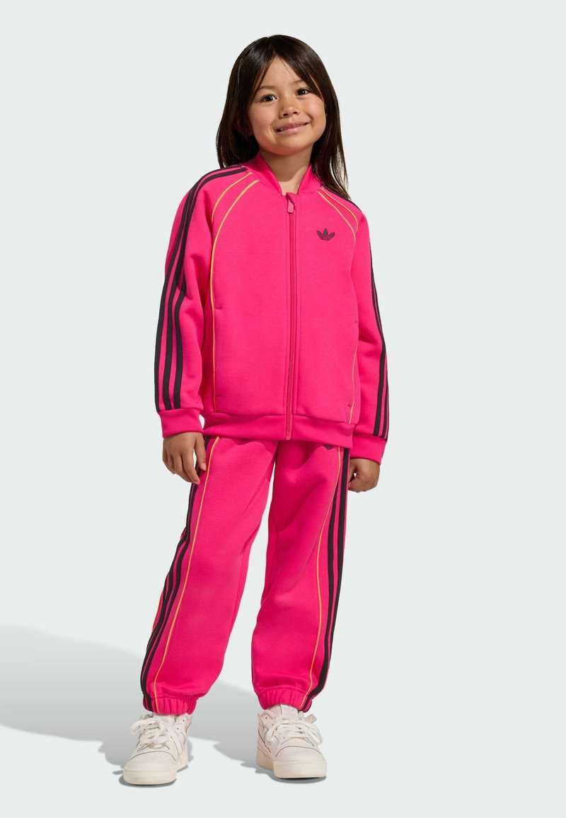 Leuchtend pinker Trainingsanzug aus weichem Stoff, bestehend aus einer Jacke mit Reißverschluss und schwarzen und gelben Streifen sowie einer Hose mit elastischen Bündchen.