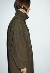 Trench coat oversize verde oliva, con colletto alto, bottoni neri e tessuto strutturato. La vista laterale mostra maniche voluminose e un fit rilassato.