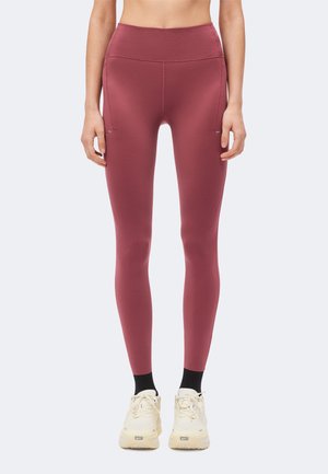 Kvinde iført mauve højtaljede leggings med sidelommer, sorte sokker og hvide Nike-sneakers stående mod en ensfarvet baggrund.