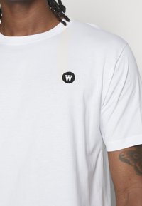 Vit bomulls t-shirt med rund hals, med en liten svart cirkulär logotyp på vänster bröstområde. Mjuk textur, korta ärmar.