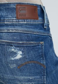 Vaqueros de mezclilla azul con detalles desgastados, que presentan una etiqueta de cuero marrón con el logo de la marca, y un pespunteado en contraste. El diseño del bolsillo trasero es visible.