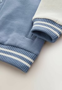 Bluza z okrągłym dekoltem w jasnoniebieskim i kremowym kolorze. Materiał polarowy z ściągaczami przy mankietach zdobionymi granatowo-białymi paskami. Zawiera boczną kieszeń.