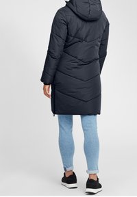 Černý péřový kabát s kapucí, zdobený vzorem v žebříčku a střední délkou. Spárován s světle modrými džíny a černými botami.