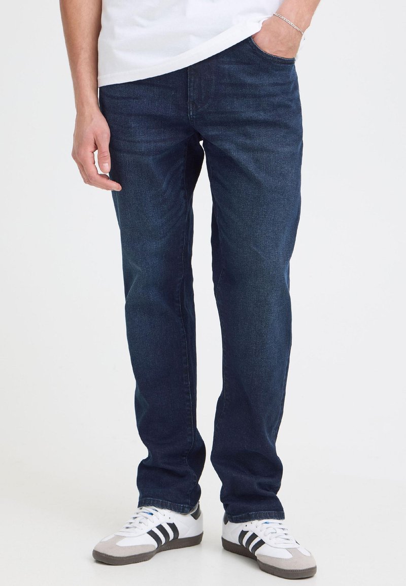 Person trägt dunkelblaue gerade Jeans, weißes T-Shirt, weiße Sneaker mit schwarzen Streifen, linke Hand in der Tasche, steht drinnen.