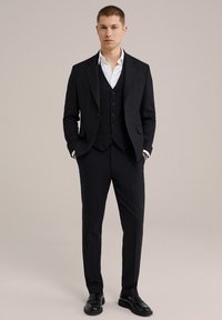 Costum format din trei piese din țesătură neagră, având un sacou ajustat, un veston cu nasturi și pantaloni croiți. Cămașă albă dedesubt, pantofi negri eleganți.