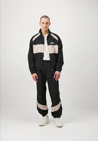 Młody mężczyzna stoi w czarno-beżowej kurtce dresowej i spodniach, z białymi sneakersami i białą koszulą, na jednolitym tle.