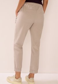 Pantalon de jogging beige clair avec taille élastique et poignets élastiques, doté de poches latérales zippées et d'une texture lisse. Porté avec des baskets beiges.