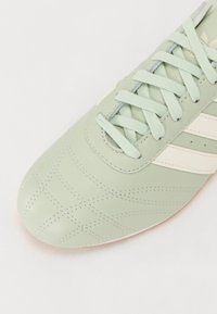 Grön lädersportssko med vita ränder, med en texturerad ovandel, platta snören och en rundad tådesign.