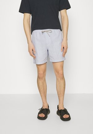 Person trägt hellblaue gestreifte Shorts mit Kordelzug, ein schwarzes kurzärmeliges Shirt und schwarze Sandalen vor einem einfarbigen weißen Hintergrund.