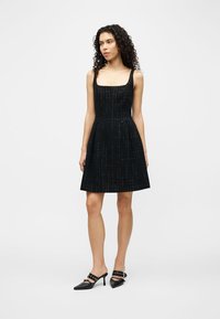 Γυναίκα στέκεται μπροστά σε απλό φόντο, φορώντας μαύρο αμάνικο φόρεμα με διακριτικό σχέδιο πλέγματος και μαύρα παπούτσια με τακούνι και μυτερή μύτη.