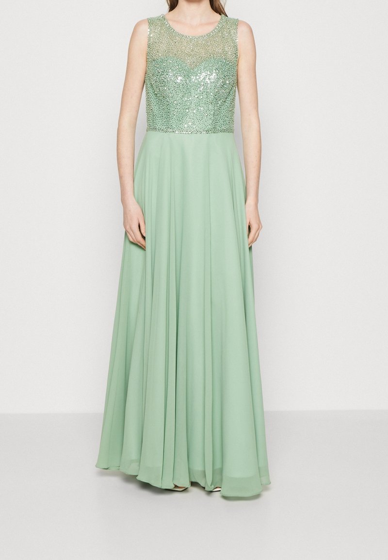 Robe de soirée vert menthe sans manches avec un corsage ajusté orné de sequins et une jupe fluide en chiffon, portée par une personne debout.