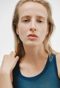 Pendientes de oro con acentos violetas. El modelo lleva una blusa sin mangas azul. El cabello suelto y ondulado, con una textura natural. Fondo minimalista.