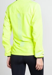 Jasně žlutá bunda s dlouhými rukávy, reflexními prvky a lehkým texturovaným materiálem; na zadní straně se nachází vertikální síťovina.