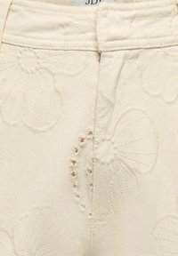 Gros plan sur un tissu crème avec des motifs floraux brodés et un détail de ceinture boutonnée.