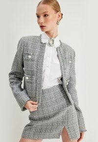 Harmaa tweed-take, jossa on purjehtimisreunat, helmi-napit ja istuva hame, jossa on sivupilkku. Valkoinen paita alla, kiiltävä rintaneula.
