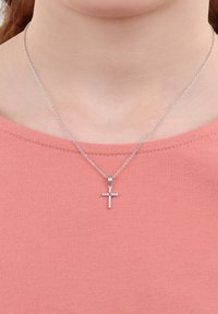 Collana a croce d'argento con piccole pietre trasparenti su una catena fine, indossata sopra una camicia rosa testurizzata.