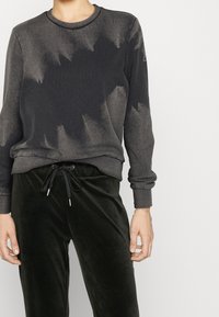 Felpa grigia scura con un sottile motivo tie-dye. Tessuto morbido e polsini a costine. Abbinata a pantaloni in velluto verde scuro con vita regolabile tramite cordino.