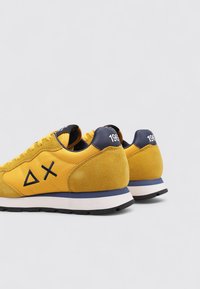 Sneaker gialle in suede e mesh, con un tallone blu navy, suola bianca e dettaglio del logo nero sul lato.