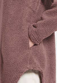 Fleecejakke i mauve med en tekstureret overflade, der har en sidelomme og en asymmetrisk kant. Lange ærmer med en afslappet pasform.