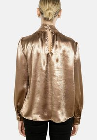 Kobieta o blond włosach związanych w kok nosi błyszczącą, brązową bluzkę z długim rękawem, z kluczowym wycięciem z tyłu i guzikiem, zestawioną z czarnymi spodniami.