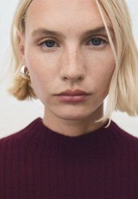 Primo piano di una persona con capelli biondi corti, che indossa un maglione a coste color bordeaux. Orecchini a cerchio in argento accentuano il look.