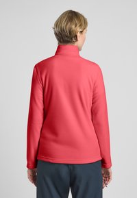 Pullover in pile rosso con mezza zip, maniche lunghe e colletto alto, caratterizzato da una texture liscia e una vestibilità casual, indossato con pantaloni scuri.