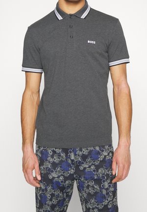 Mann trägt ein dunkelgraues Poloshirt mit weiß gestreiftem Kragen und Ärmeln sowie gemusterte blaue und graue Hosen mit abstraktem Blumendesign.