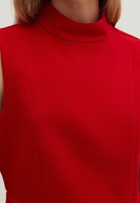 Mujer con una blusa roja sin mangas y un cuello alto, que muestra el escote y la parte superior del pecho, contra un fondo liso.
