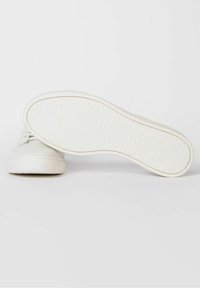 Bílé tenisky s hladkým koženým svrškem, kulatou špičkou a texturovanou gumovou podrážkou s kruhovými vzory. Šňůrky jsou výrazné.