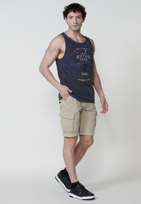 Námořnický tílko s grafikou "Motor Club", béžové cargo šortky s několika kapsami a černé sportovní boty s bílou podešví.