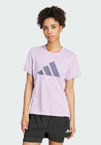 Vaalean violetti urheilupaita, jossa on suuri harmaa Adidas-logo. Paidassa on lyhyet hihat ja pyöreä kaula-aukko, ja se on valmistettu hengittävästä kankaasta.