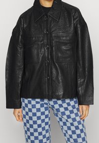 Sort læderjakke med struktureret overflade, oversized pasform, store forlommer og knaplukning, parret med blårutede bukser.