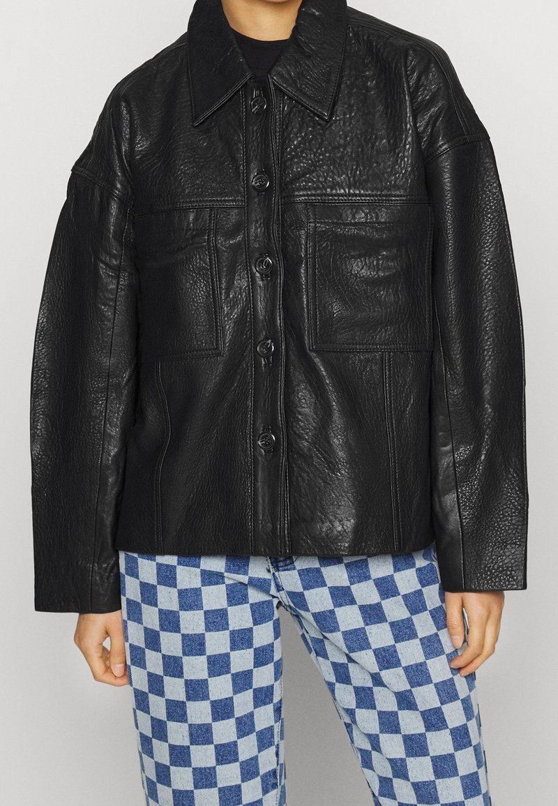 Sort læderjakke med struktureret overflade, oversized pasform, store forlommer og knaplukning, parret med blårutede bukser.