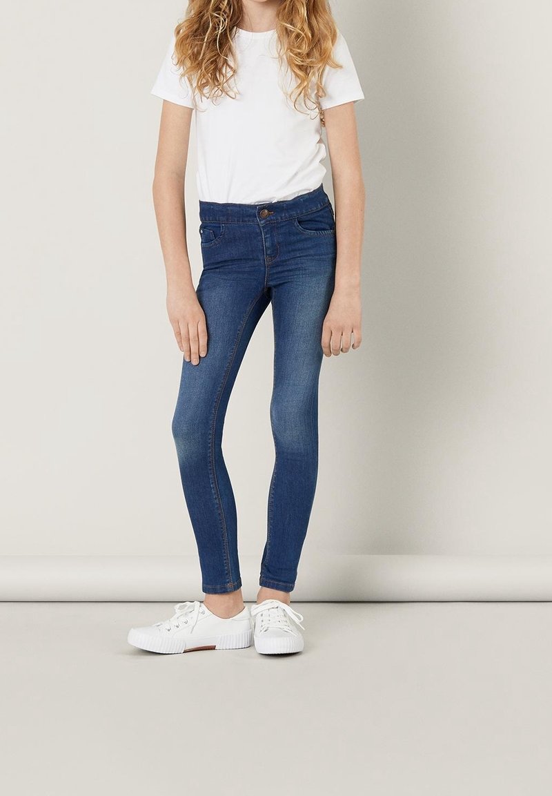 Enfant aux longs cheveux bouclés, debout, portant un t-shirt blanc, un jean skinny bleu et des baskets blanches à lacets, sur fond uni.