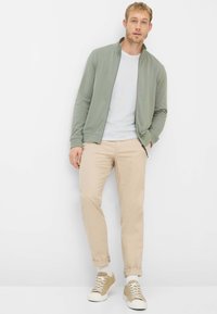 Lichtgroene jas met rits over een wit T-shirt, gecombineerd met beige broek en tankleurige sneakers. Gladde stof, moderne pasvorm, casual ontwerp.