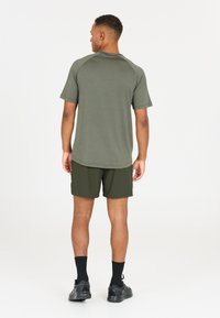 Olivgrön t-shirt med kort ärm och raglanärmar, kombinerad med mörkgröna shorts; modellen bär svarta strumpor och svarta sportskor.