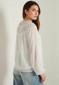 Blusa bianca a maniche lunghe realizzata in tessuto trasparente con pois testurizzati, dotata di scollatura arricciata e vestibilità ampia. Abbinata a jeans azzurri.