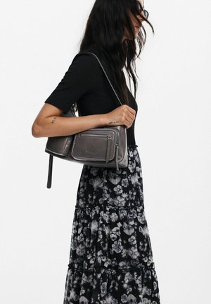 Sac à main en cuir gris avec plusieurs poches zippées, un design épuré et une longue bandoulière. Le mannequin porte un haut noir et une jupe à motifs floraux.