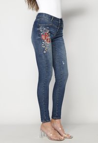 Mörkblå skinny jeans i denim med blommig broderi på vänster lår, subtil slitage och en standard midja. Klara blockklackade sandaler.