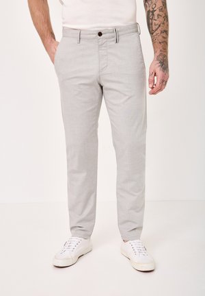 Man wearing light gray slim-fit trousers, white sneakers, and a white shirt, standing with one hand in pocket.