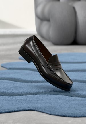 Black leather loafer with moccasin stitching displayed on blue textured carpet, with gray tufted furniture in the blurred background.