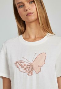 Camiseta blanca de algodón con un gran diseño de mariposa bordada en rosa en el frente, con detalles intrincados y acentos texturizados.