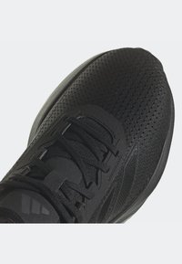 Gros plan sur une chaussure de sport noire avec une tige en maille, des lacets noirs et un logo de marque discret sur la languette et sur le côté.