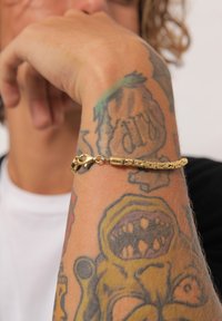 Bracelet chaîne en or avec un design de maillon complexe ; surface texturée ; doté d'un fermoir robuste. Porté sur un bras tatoué contre un fond uni.