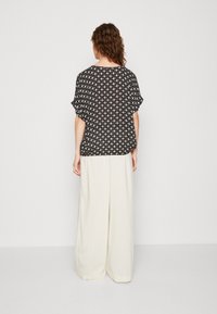 Schwarze gemusterte Bluse mit kurzen Ärmeln, kombiniert mit weißen, gestreiften, weit geschnittenen Hosen. Die Bluse hat ein geometrisches Design.