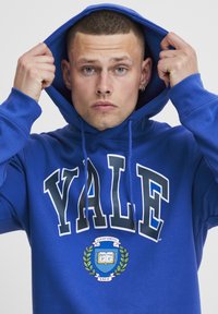 Felpa blu con la scritta "YALE" in evidenza. Presenta un emblema decorativo con foglie di alloro e testo. I lacci sono visibili sul cappuccio.
