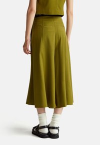 Persoon in olijfgroene, wijde, cropped broeken, met witte sokken en zwarte sandalen, gezien vanuit de achterkant tegen een effen achtergrond.