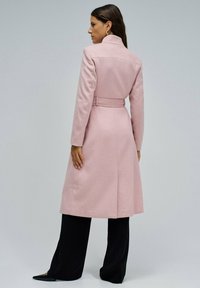 Duga ružičasta kaput od vune s pojačalom s pojasom, krojenim ovratnikom i dugim rukavima; nosi se preko crnih hlača širokih nogavica.