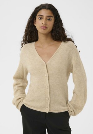 Beige wool cardigan with a V-neck, long sleeves, and button closure. Soft texture, loose fit, and ribbed hem detail. Paired with black pants.